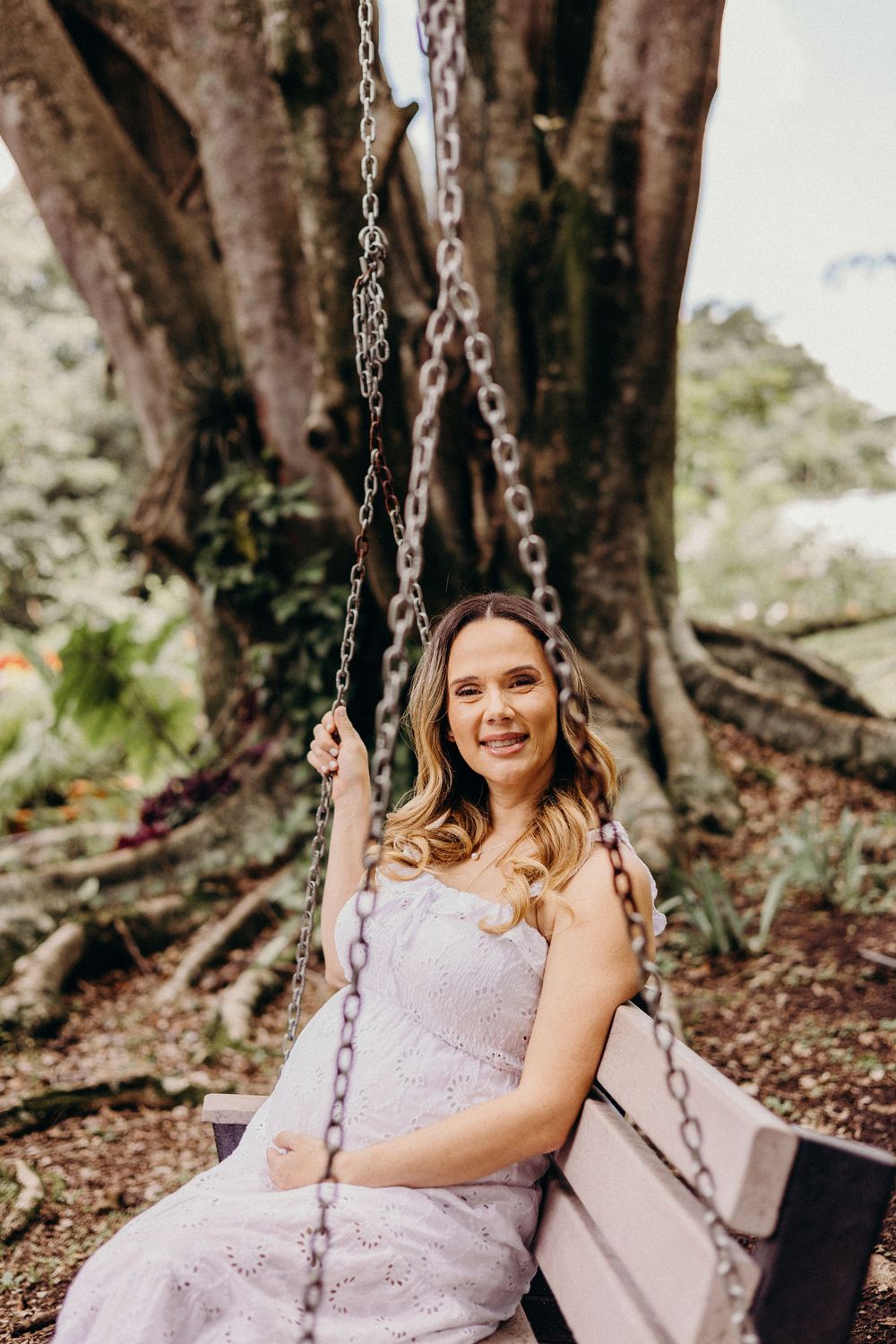 A person in a lavender dress sits on a chain swing under large trees in a natural outdoor setting.