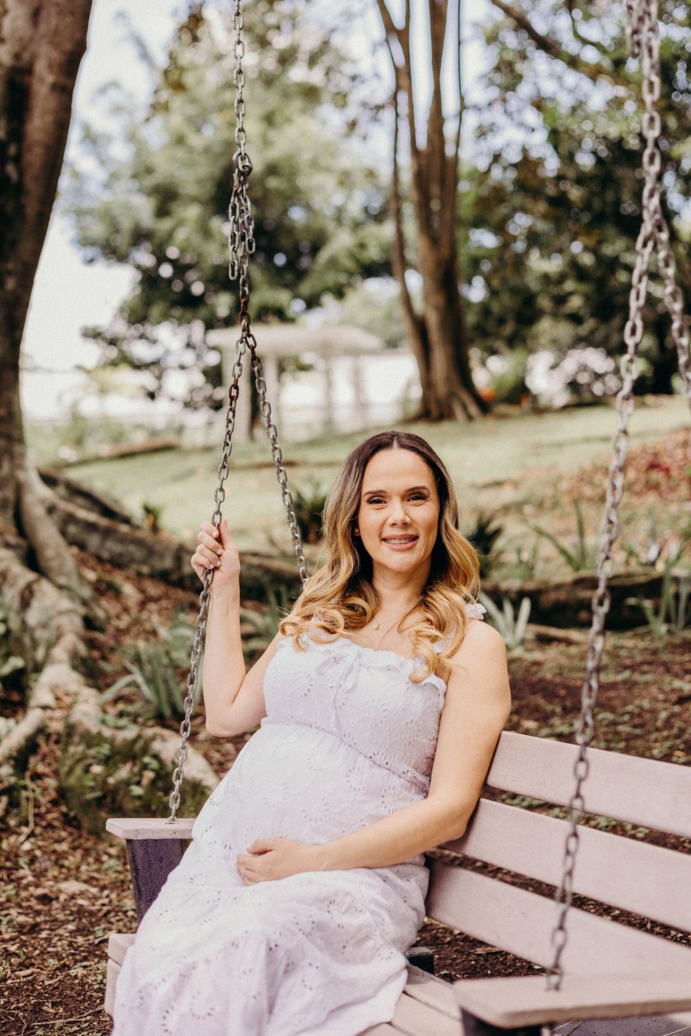 A person in a light pink dress sits on a wooden swing in a serene outdoor garden setting.
