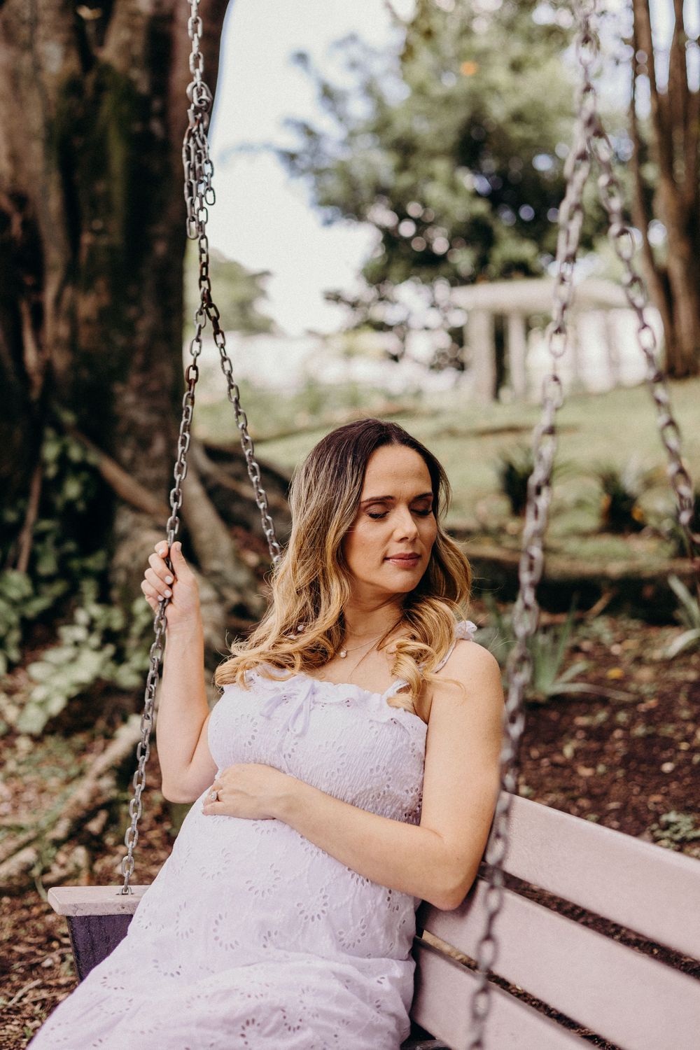 A person in a lavender lace dress sits peacefully on a wooden swing in a sunlit garden setting.