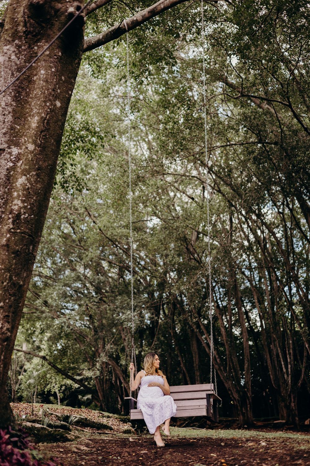 A woman in a white dress sits on a wooden swing in a serene forest setting with dappled sunlight.