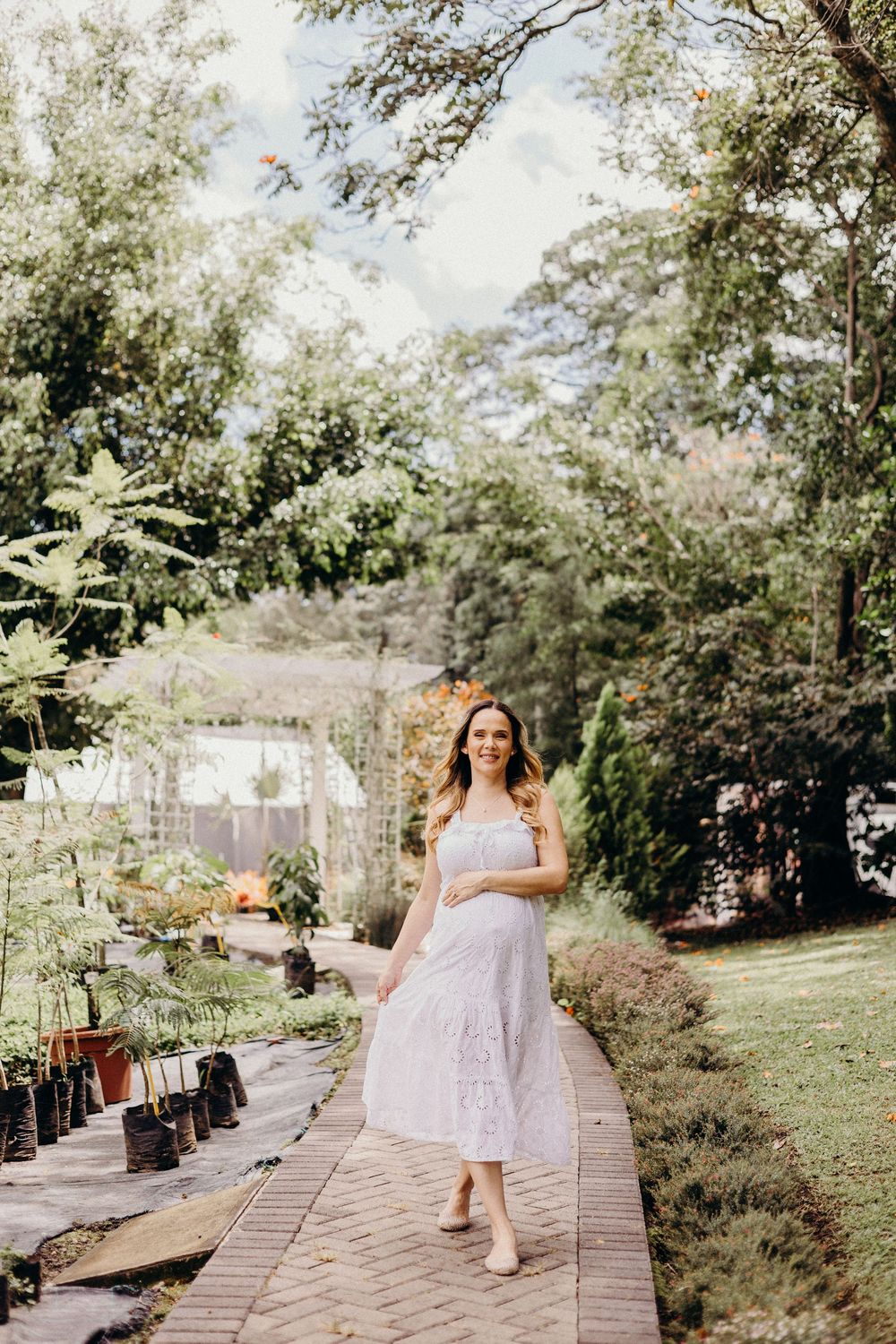 A woman in a white sundress walks along a garden path surrounded by lush greenery and trees.