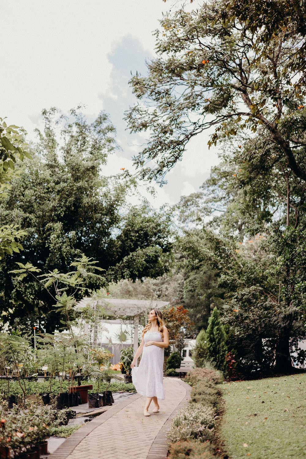 A person in a flowing white dress walks along a stone pathway in a lush garden setting on a sunny day.
