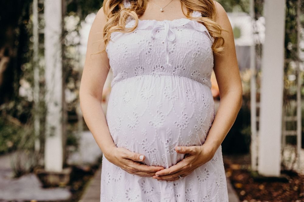 A pregnant woman in a lavender lace maternity dress poses in front of a white picket fence during a photo session.