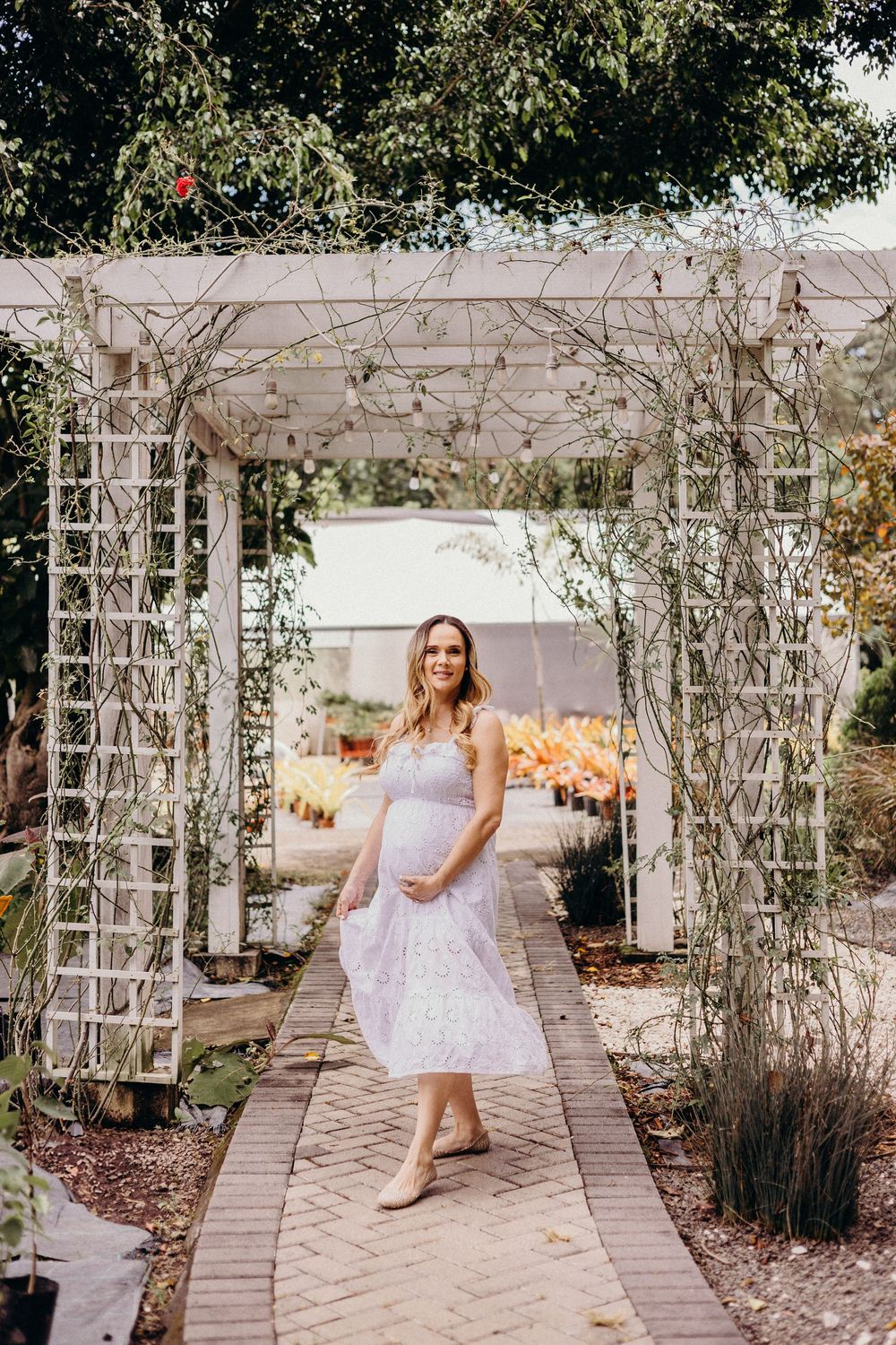 A bride in a white lace dress poses playfully in a garden pergola with climbing vines and stone pathway.