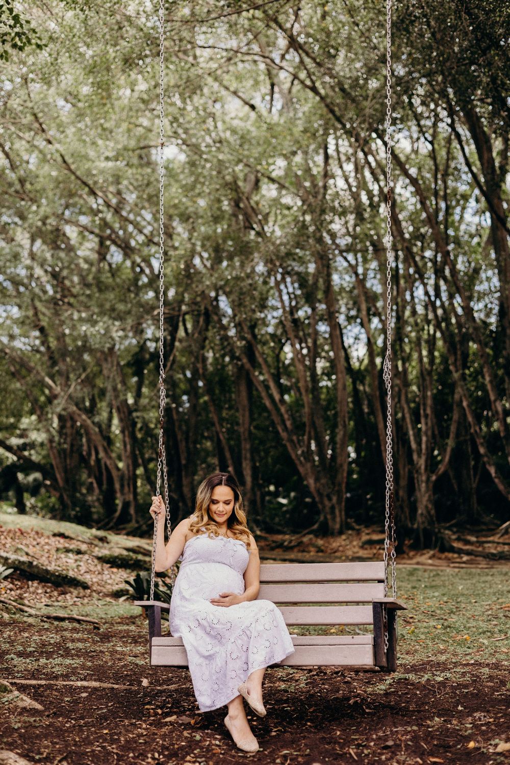 A dreamy outdoor photoshoot in a wooded area with someone sitting on a wooden bench wearing a flowy white dress.