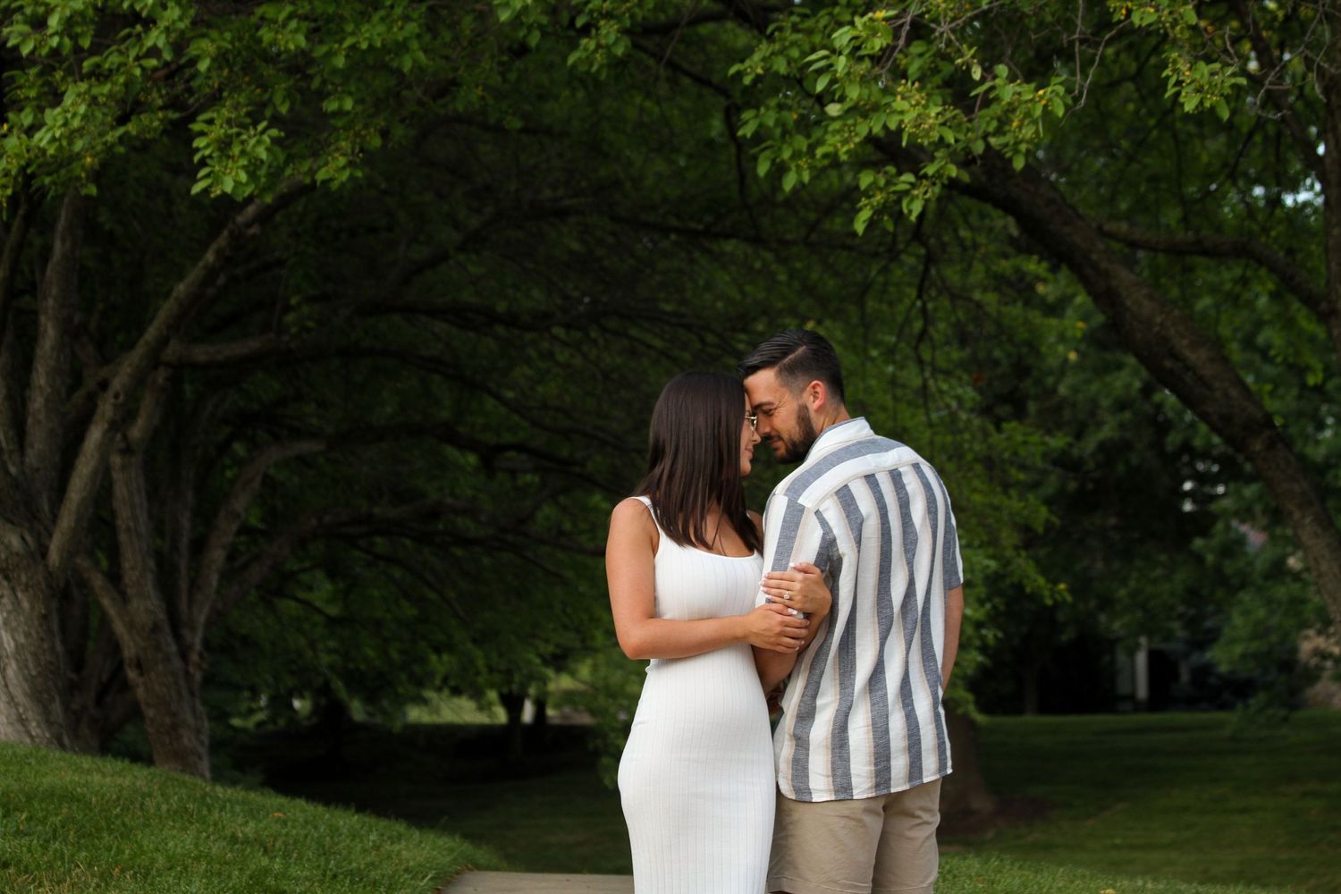 A man and woman lean their foreheads together under a dramatic canopy of leafy green trees during an engagement session.
