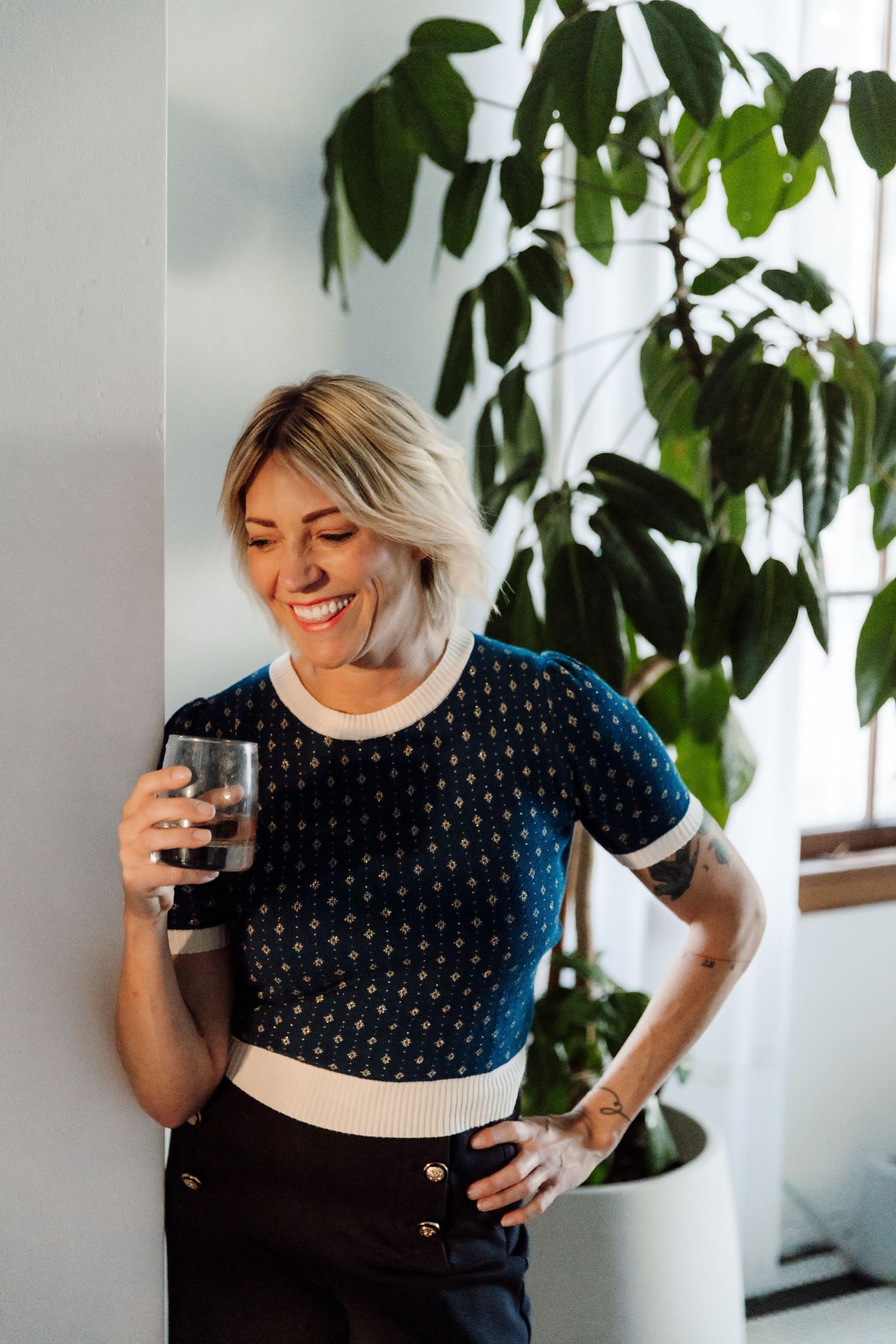 A person in a navy polka dot sweater smiles and holds a coffee mug while standing near indoor plants.