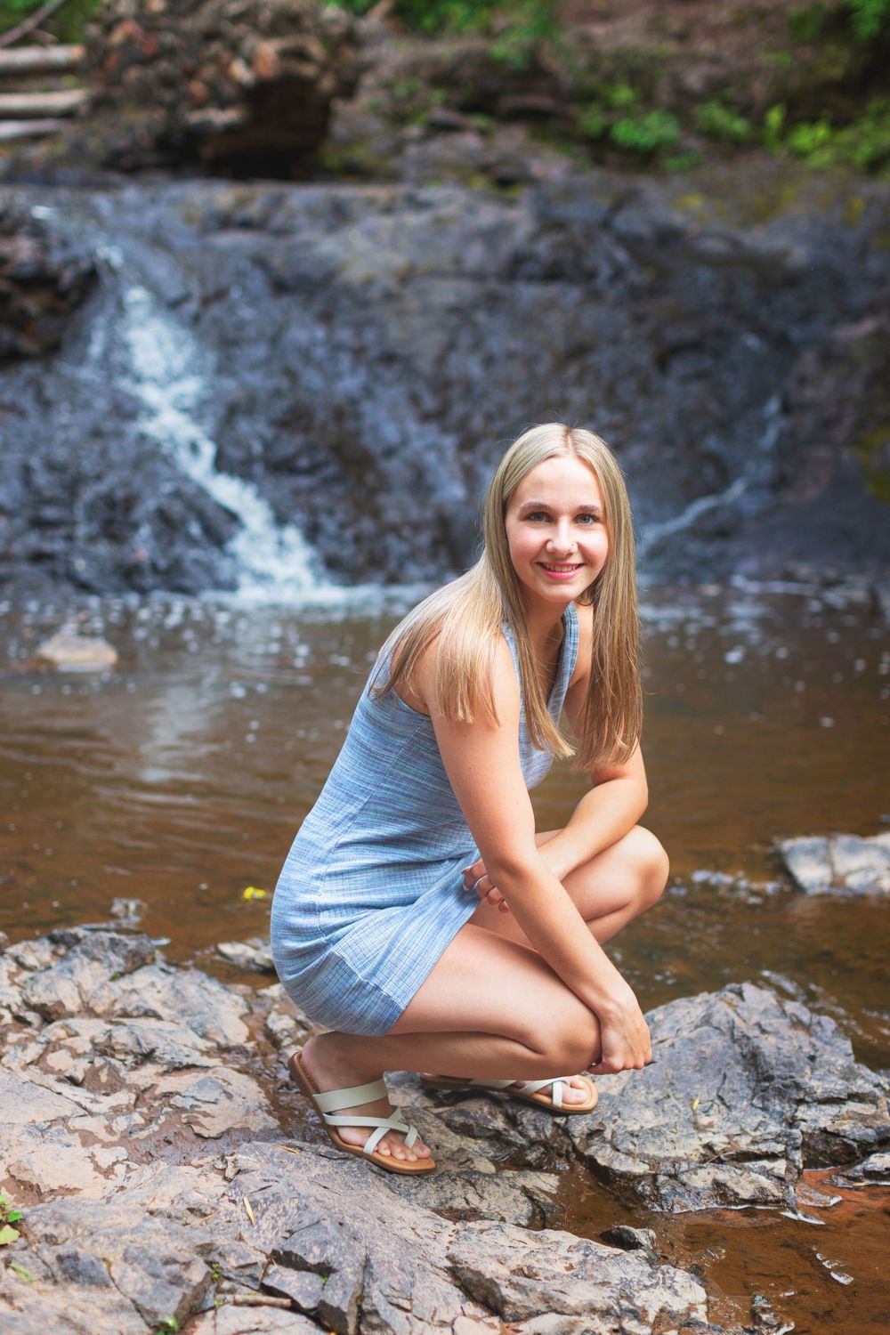 A person in a blue dress poses on rocks near a flowing stream in a natural outdoor setting.