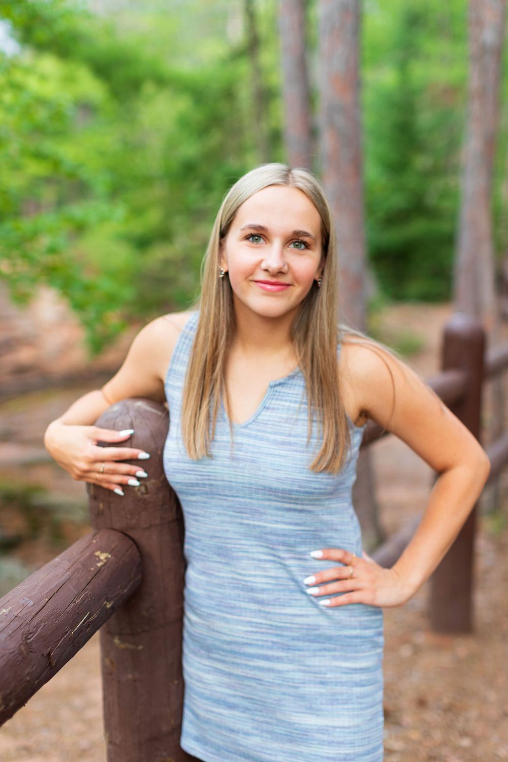 A person in a light blue sleeveless top poses casually against a wooden railing in an outdoor wooded setting.