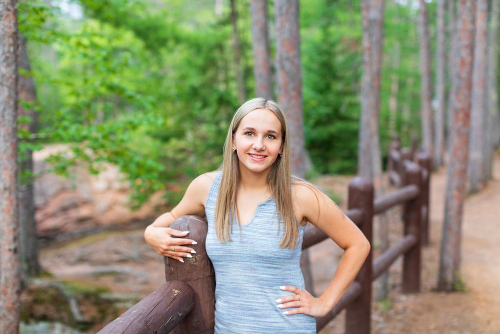 A person in a blue tank top poses outdoors along a wooden boardwalk in a lush forest setting.
