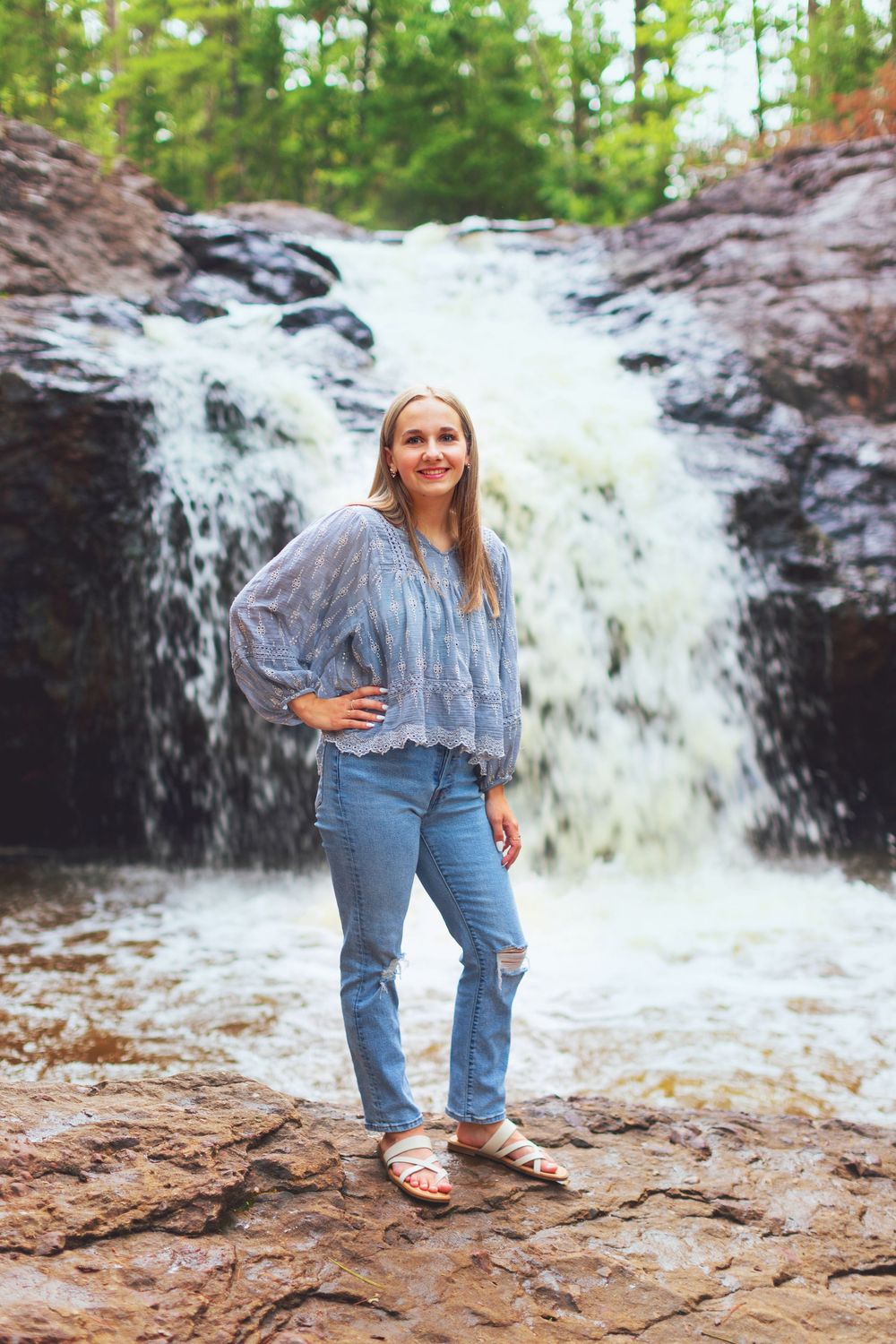 Person in denim poses near a rushing waterfall against rocky terrain in a nature setting.