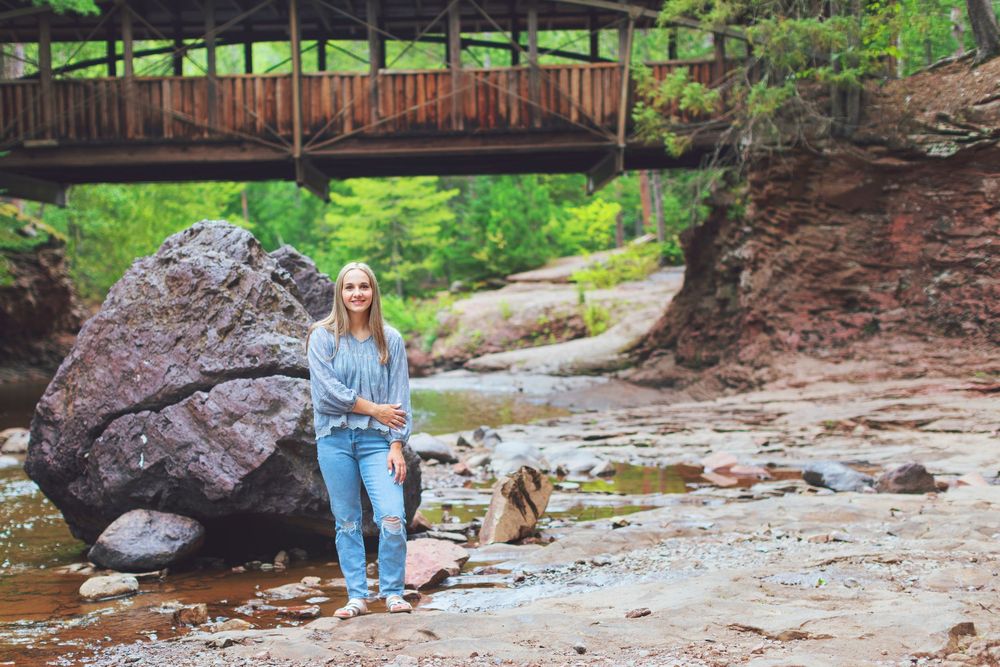 A person crouches near a flowing stream surrounded by lush greenery and rocks in a natural outdoor setting.