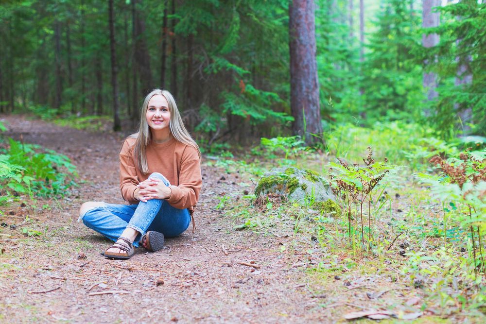 Someone in an orange sweater sits cross-legged on a forest trail surrounded by lush green trees.