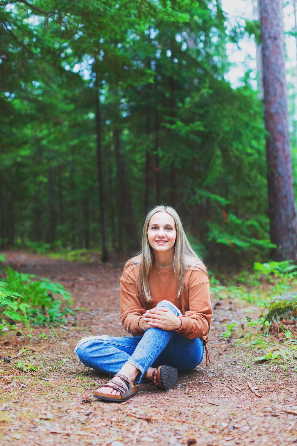 Someone wearing an orange sweater and jeans sits casually on a pine needle covered forest floor.