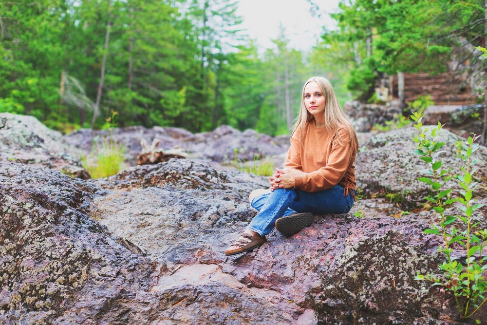 Someone in an orange sweater sits on rocky terrain in a verdant forest setting.
