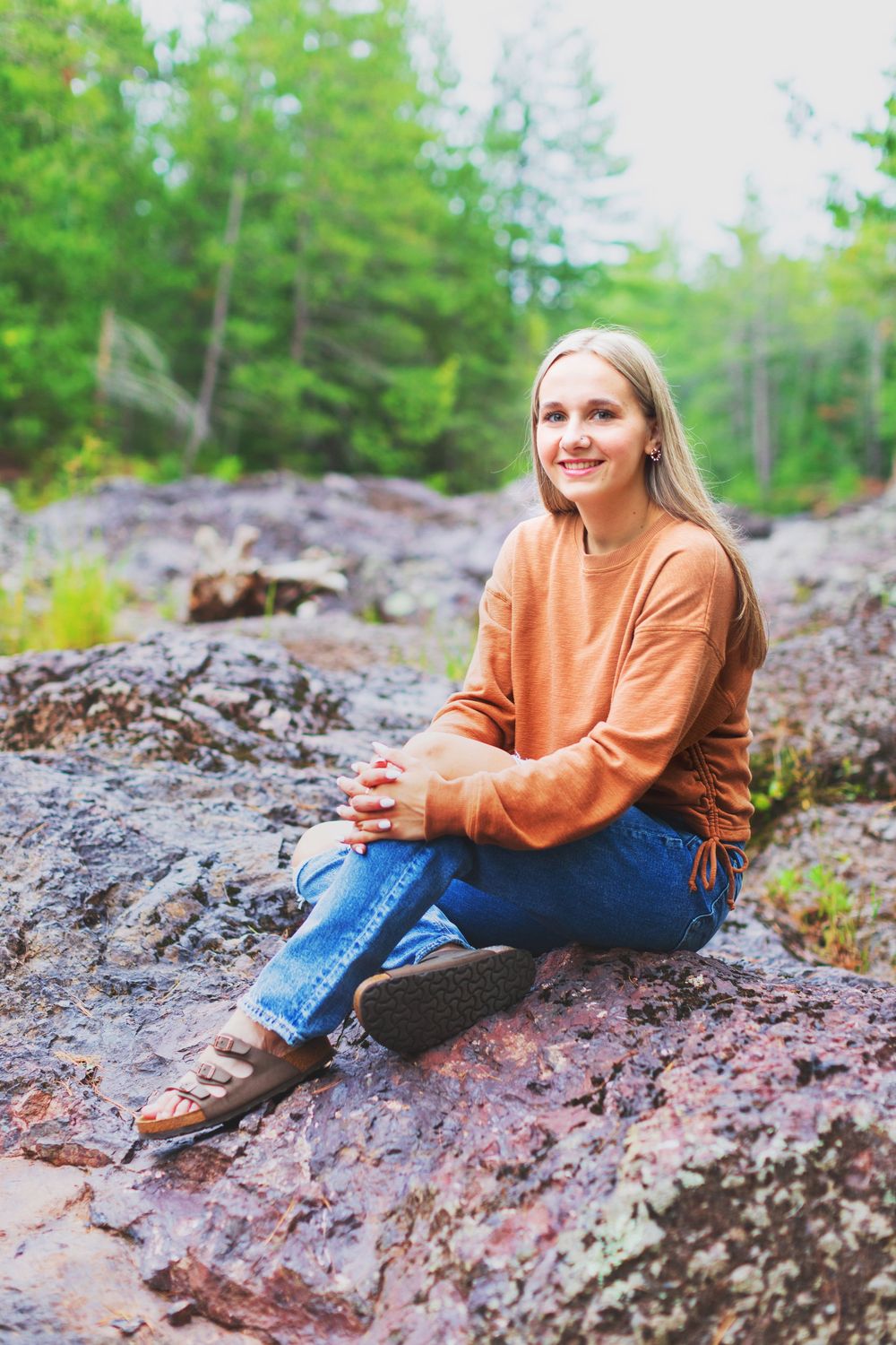 Person in orange sweater and jeans sits casually on rocky terrain surrounded by green forest vegetation.