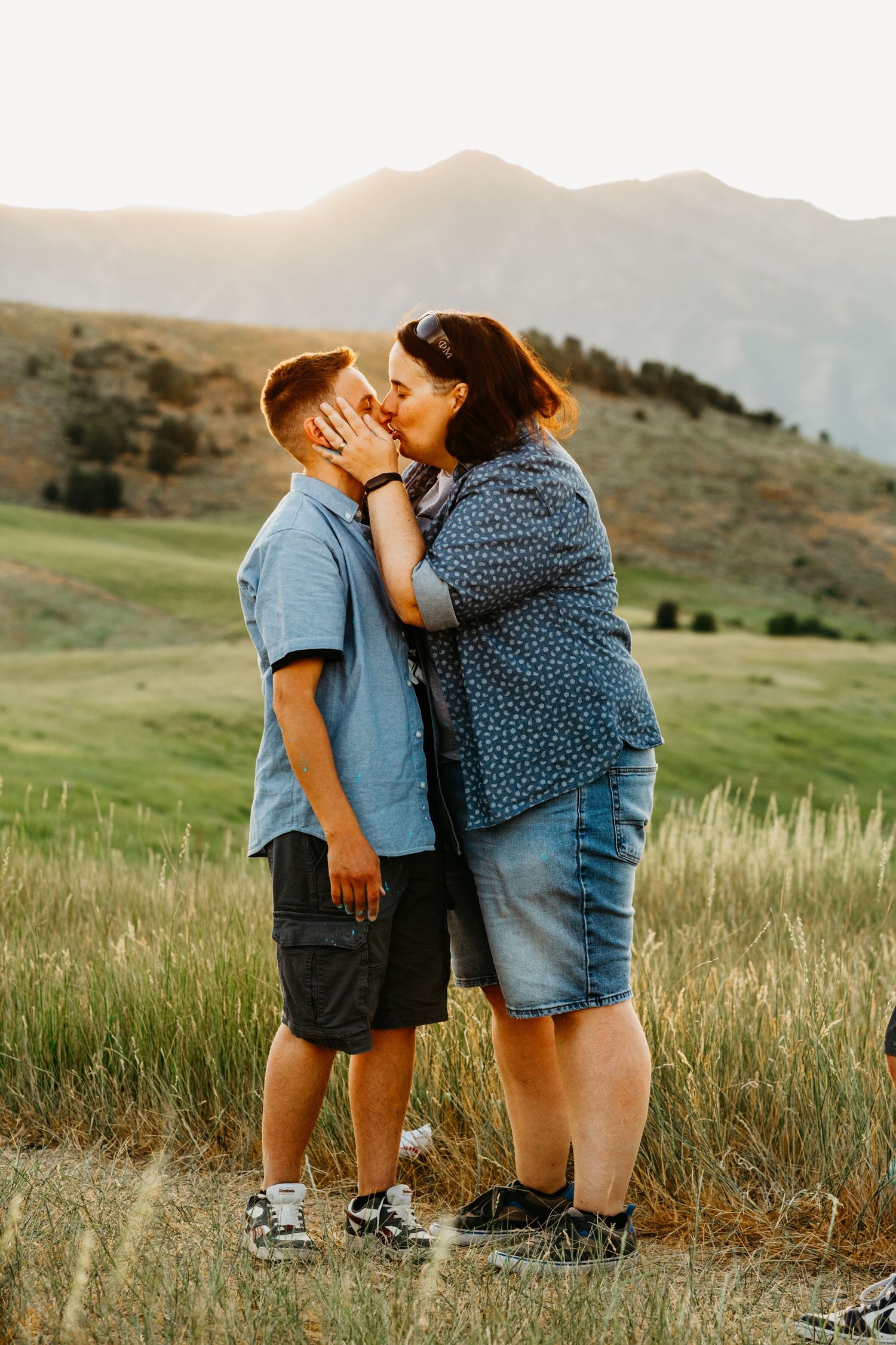 Two people share an intimate moment in a grassy field with mountains in the background during golden hour.