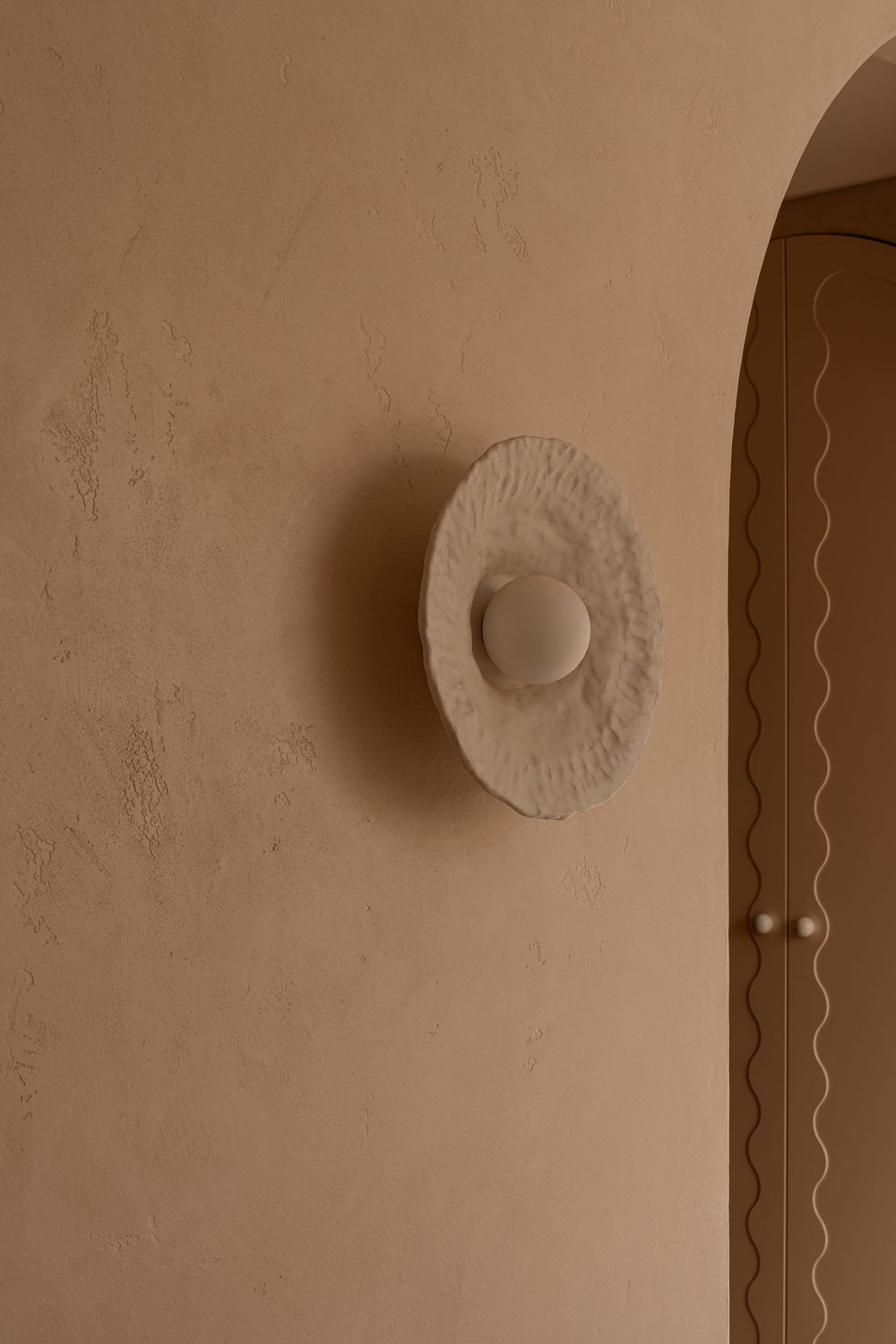 A circular white plaster wall sconce mounted on a beige wall next to an arched doorway with decorative trim.