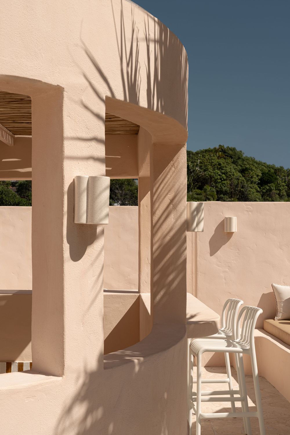 White plastic chairs line a modern Mediterranean-style terrace with geometric shadows cast on cream-colored walls.