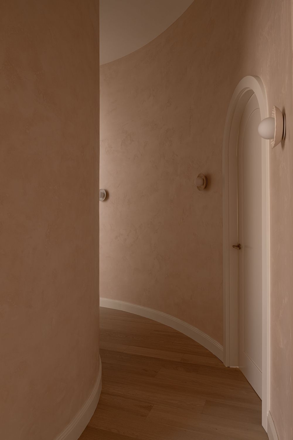 A long empty hallway with beige walls, white trim, wall sconces, and hardwood flooring extends into the distance.