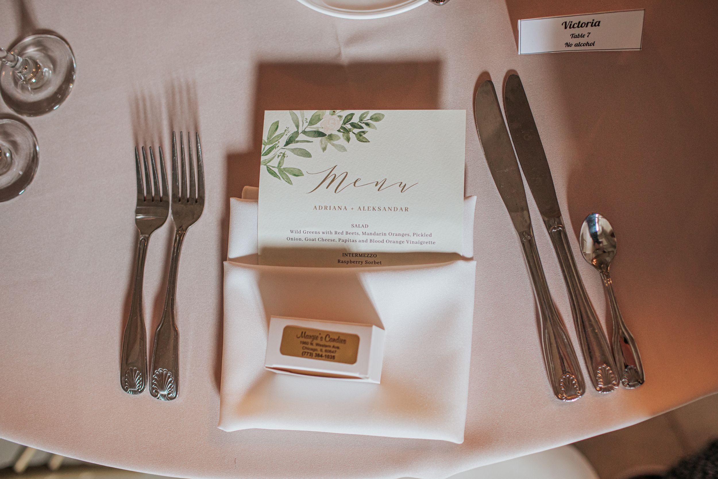 An elegant place setting with silverware, white napkin, and botanical menu card on a blush pink tablecloth.