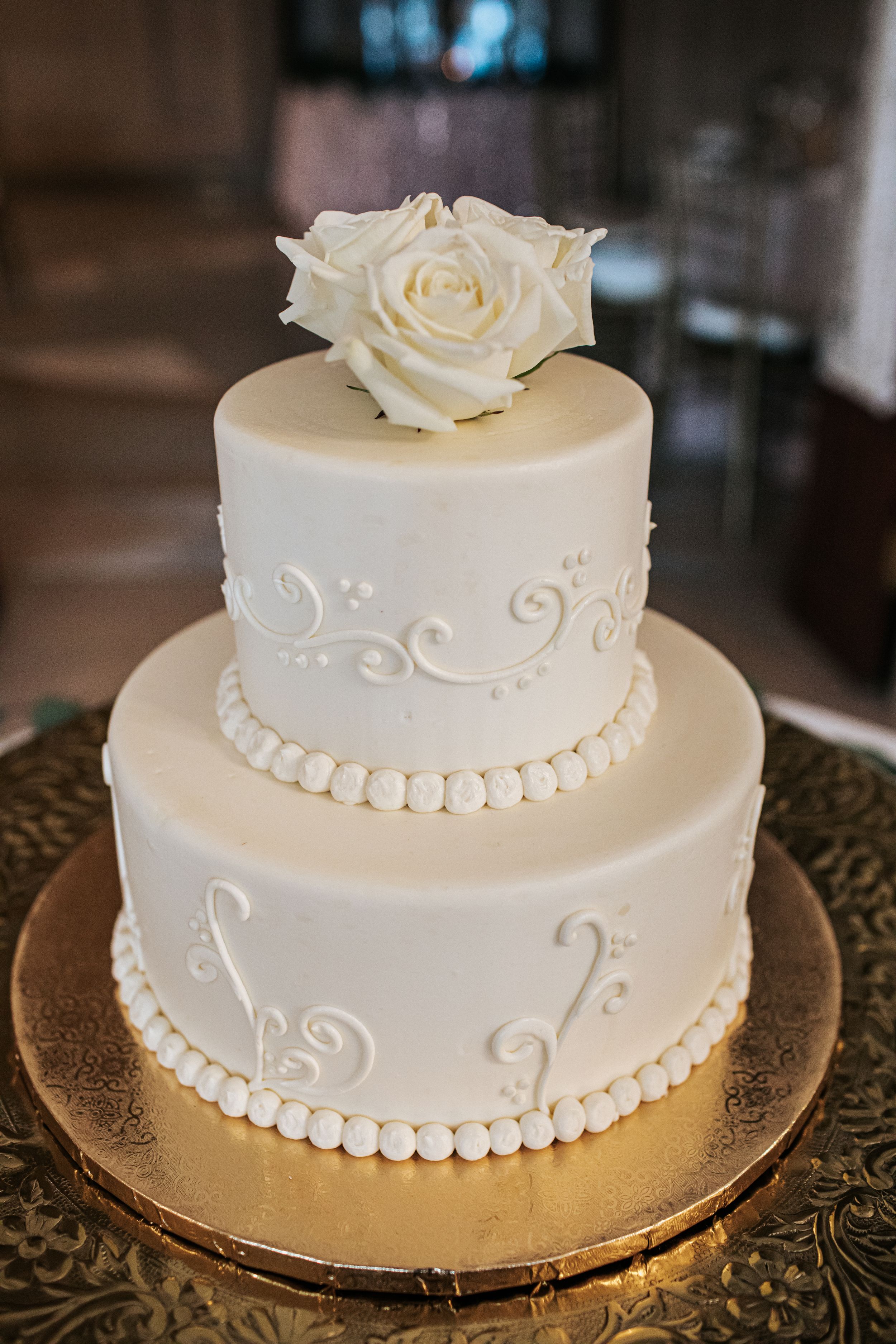 A simple two-tiered white wedding cake with delicate swirl designs and pearl border decorations topped with a white rose.