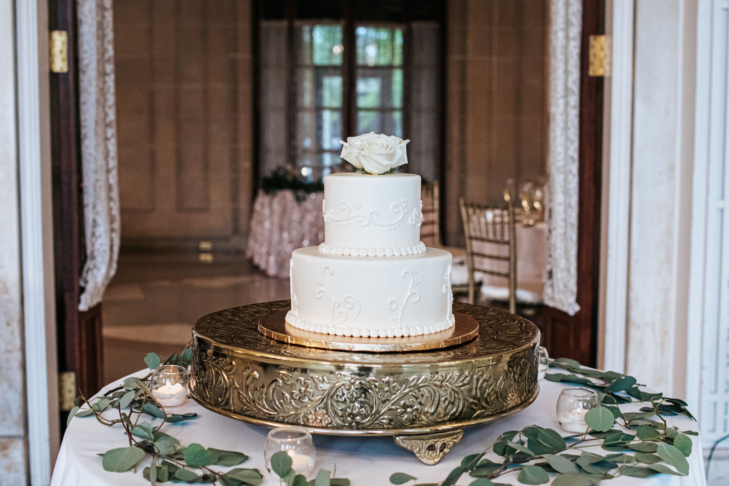 Elegant white two-tier wedding cake displayed on ornate gold stand with eucalyptus leaves and lace curtains in background.