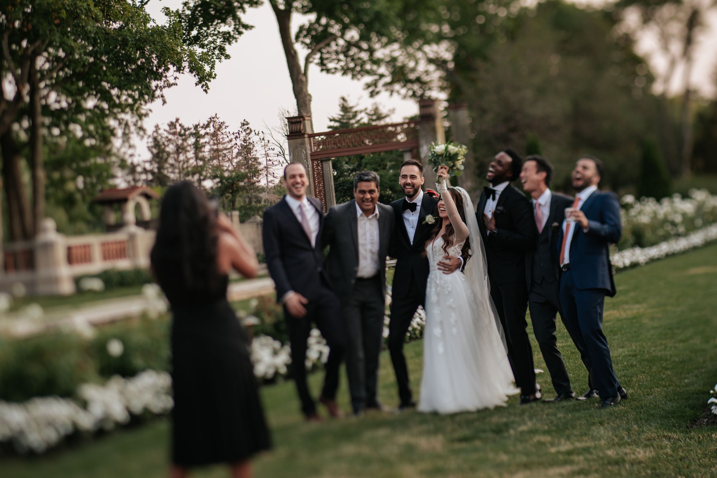 Wedding party stands together on a lush green lawn during an outdoor garden ceremony at sunset.