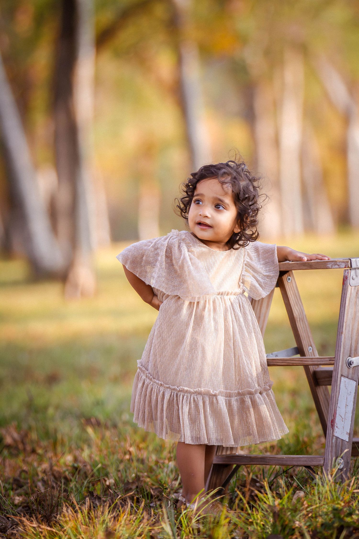 A toddler in a white vintage dress explores an outdoor setting with fallen autumn leaves and wooden chairs.