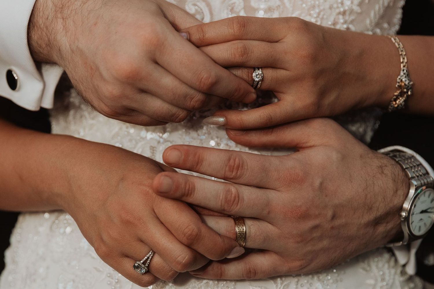 Close-up of bride and groom hands showing wedding rings, diamond engagement ring, and gold watch.