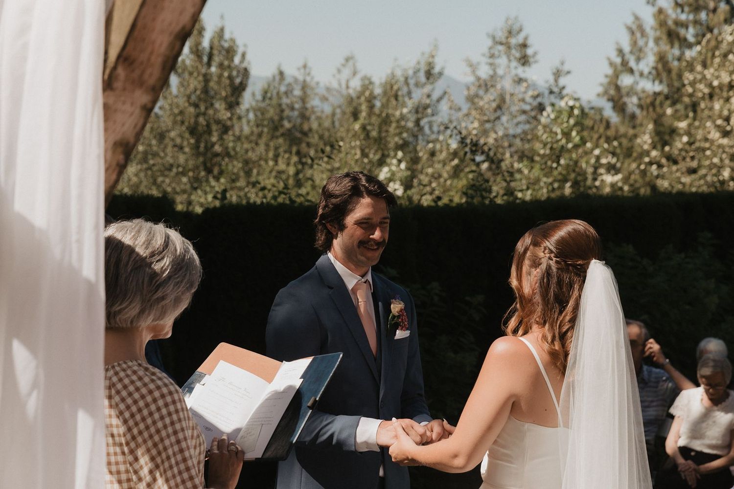 Groom smiles lovingly at bride while holding hands during intimate outdoor wedding ceremony.