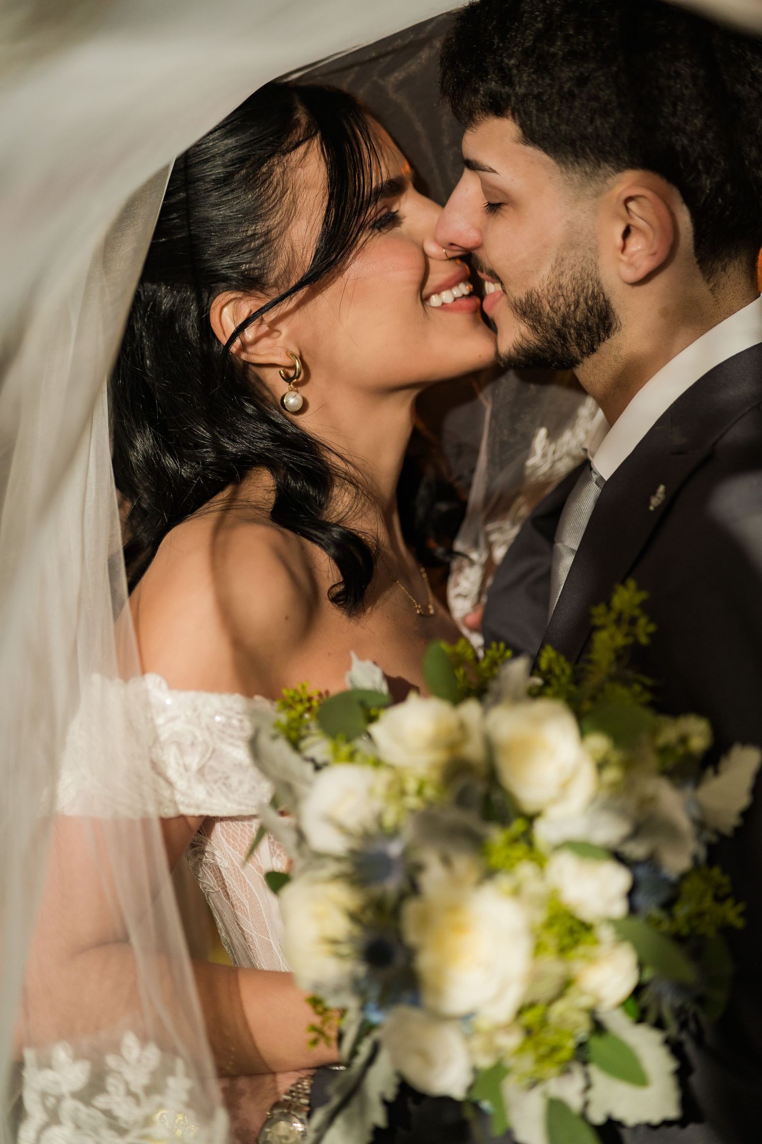 A romantic wedding couple shares an intimate kiss while holding a white floral bouquet against a light background.