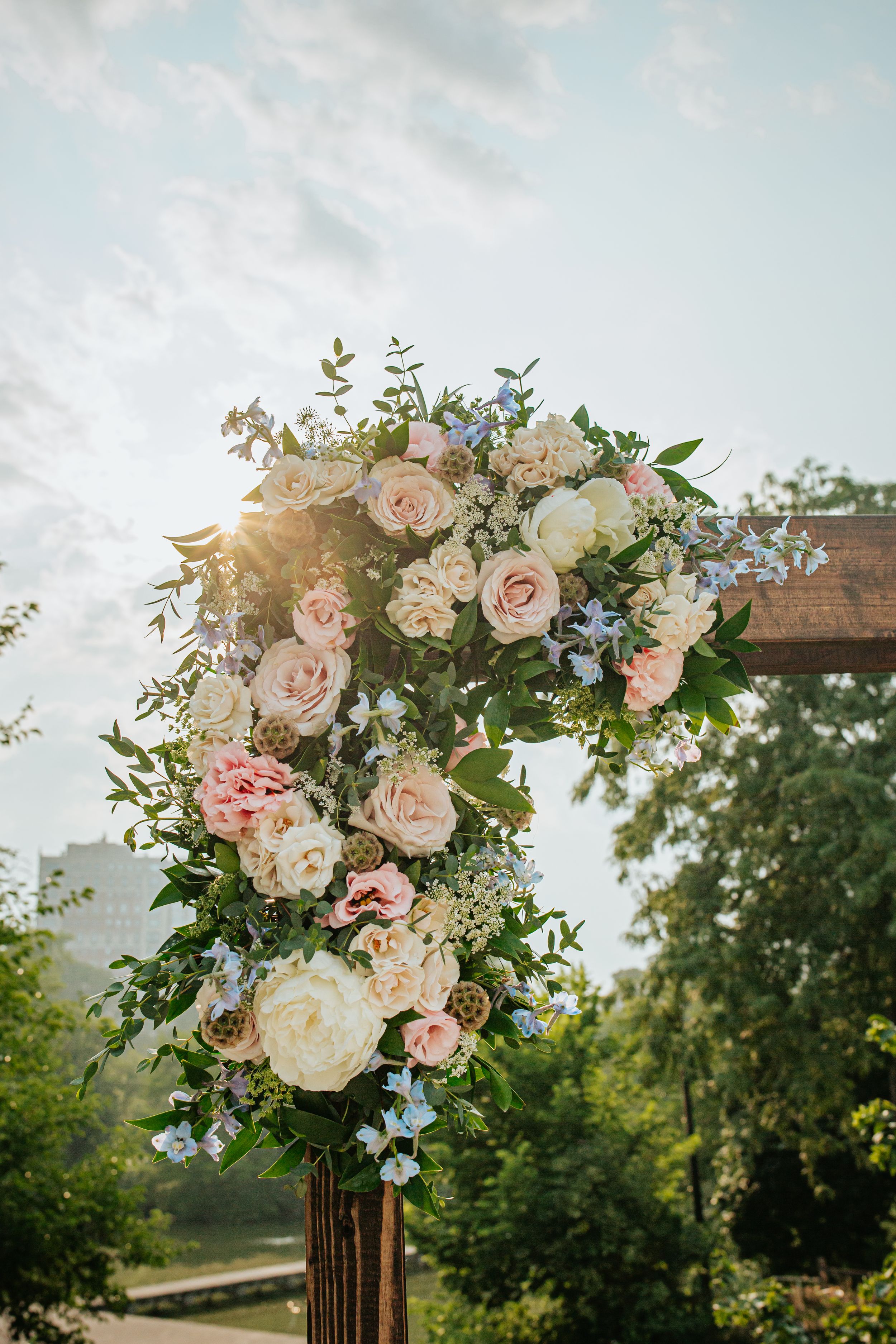 Beautiful floral arch decoration with pink and white roses and delicate blue flowers against a sunlit outdoor backdrop.