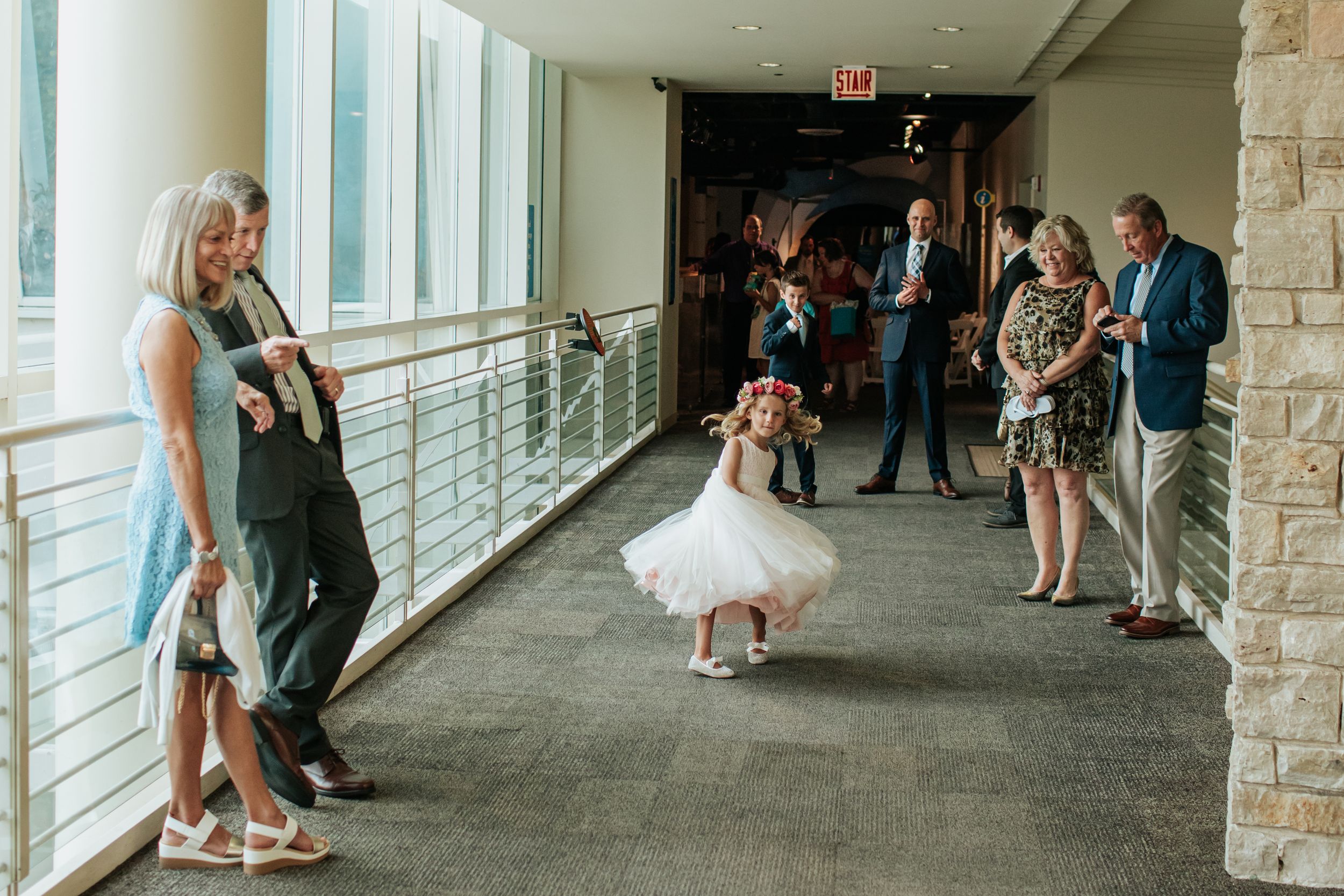 Wedding guests line a bright hallway with glass windows and stone walls as they wait for the ceremony to begin.