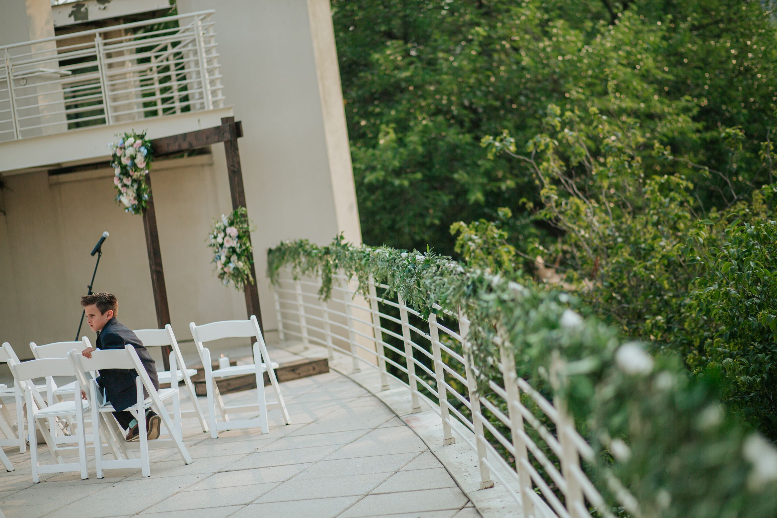 A white rocking chair sits on a wraparound porch surrounded by lush green foliage and trees.