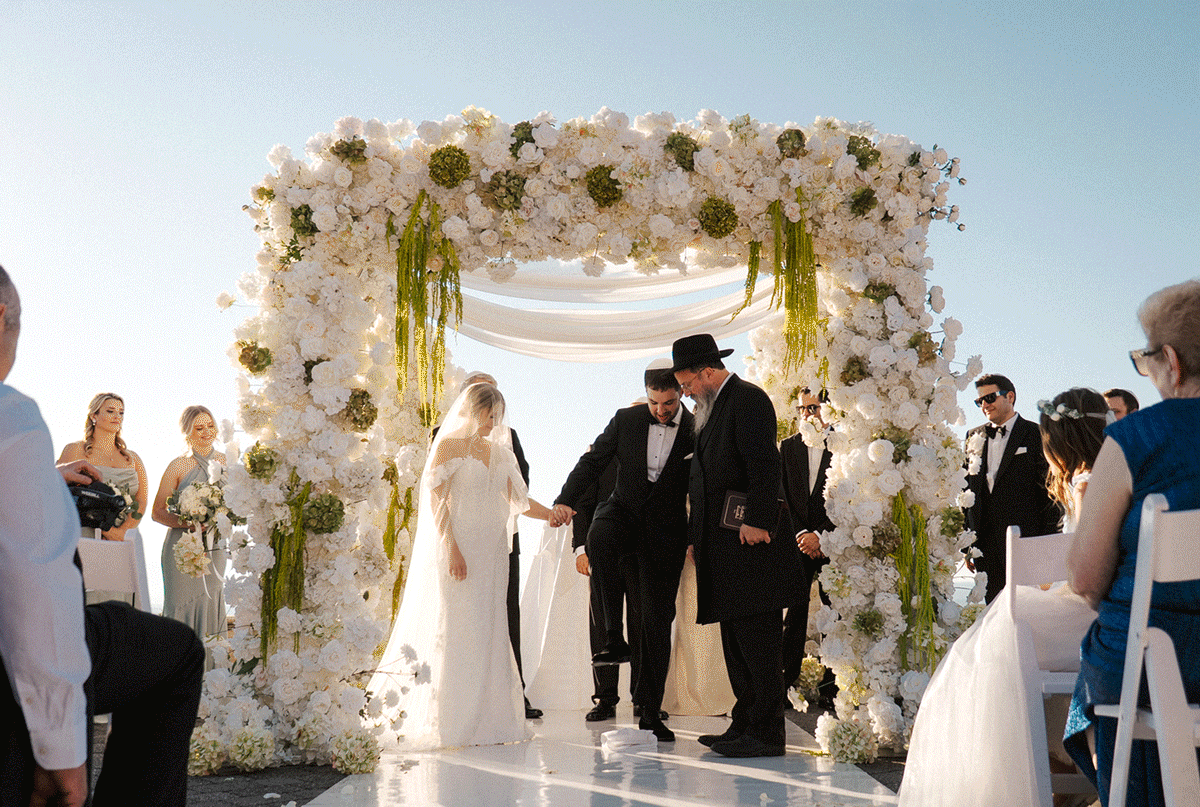 A wedding ceremony takes place under a white floral arch at an elegant outdoor venue with guests in attendance.