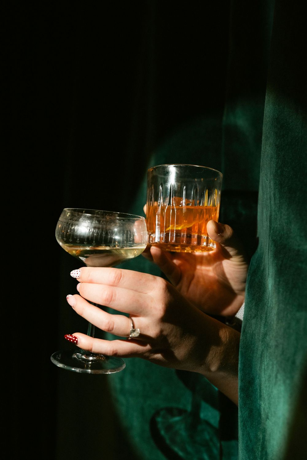 Two hands holding wine glasses in a dimly lit setting with a green background.
