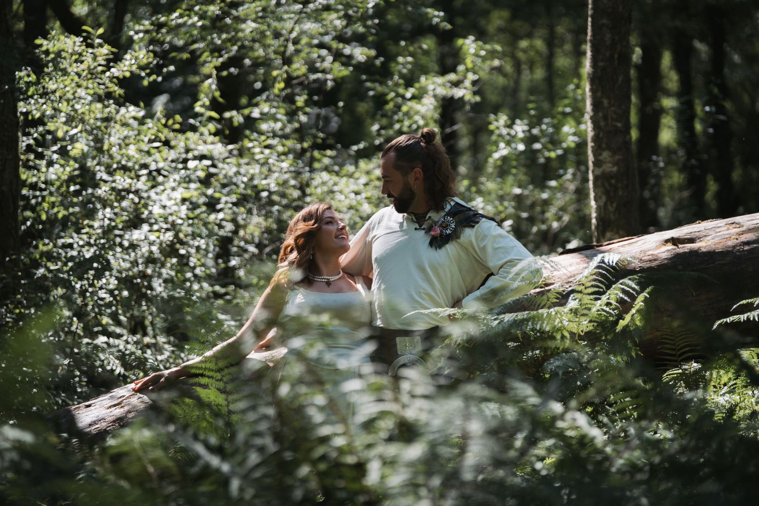 A couple poses outdoors in wedding attire, with the groom wearing a traditional Scottish kilt and the bride in a lace dress.