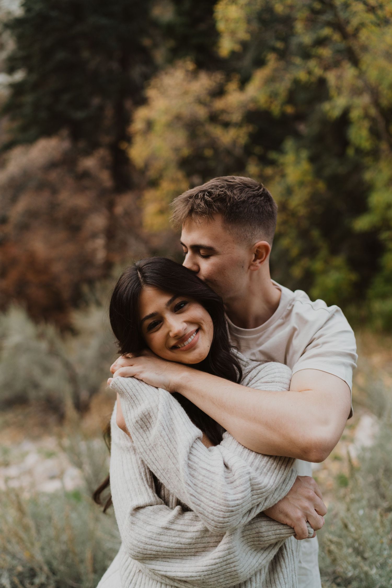 Couple embracing in casual outfits against blurred autumn foliage background.