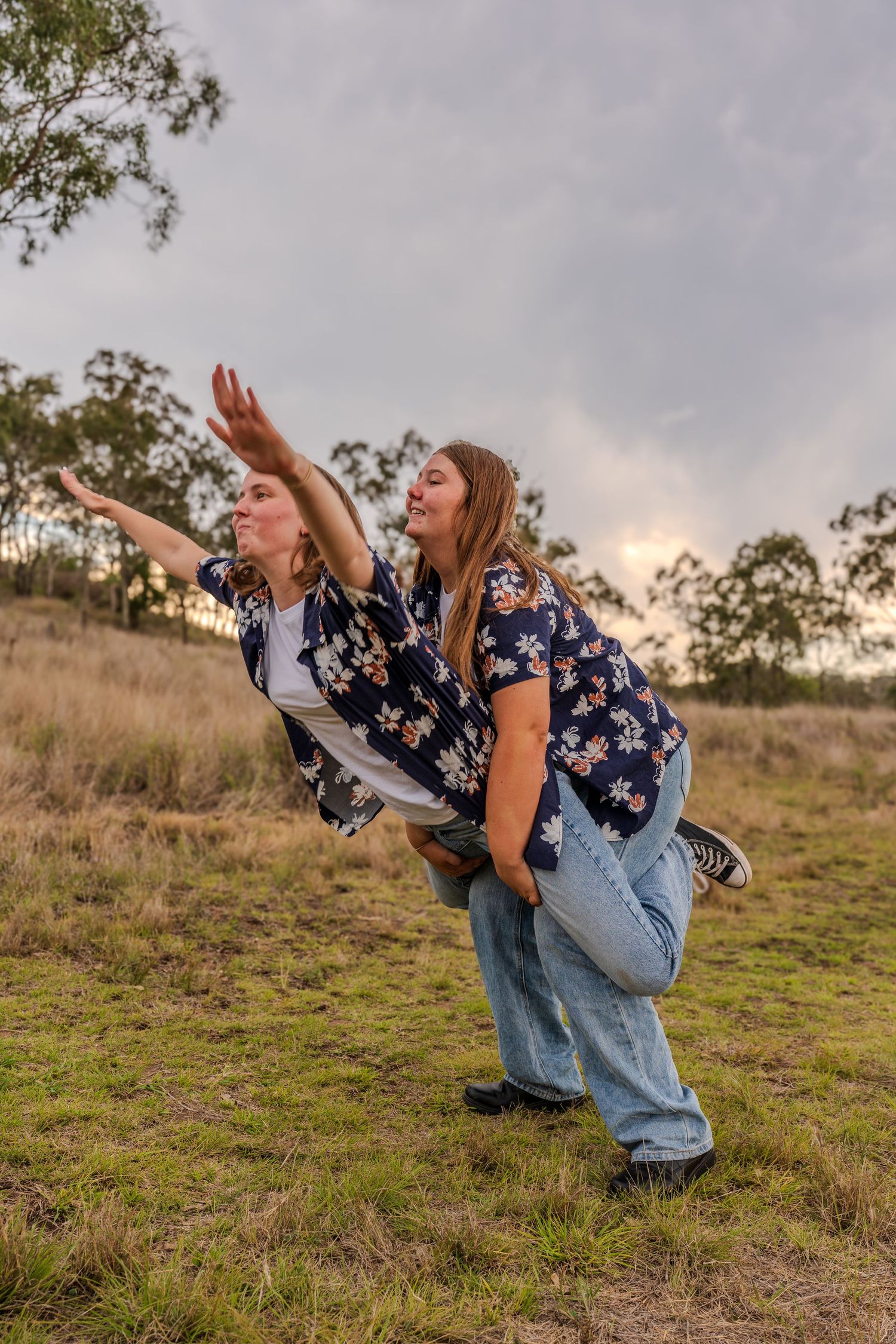 Two people create a playful airplane pose on a grassy hill at sunset.
