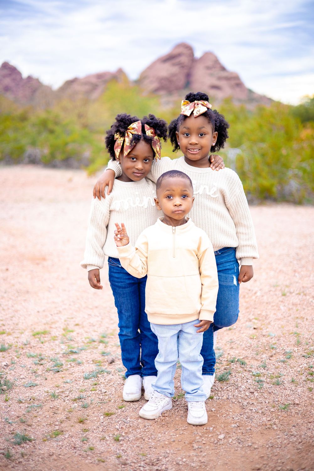 Three children in matching cream sweaters and jeans pose together on rocky desert terrain with mountains in background.