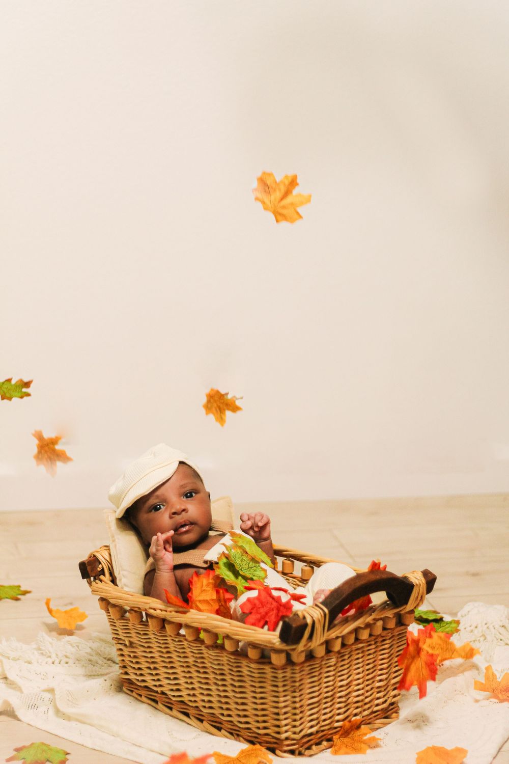 Baby in autumn-themed photo with falling orange leaves and wicker basket setup.