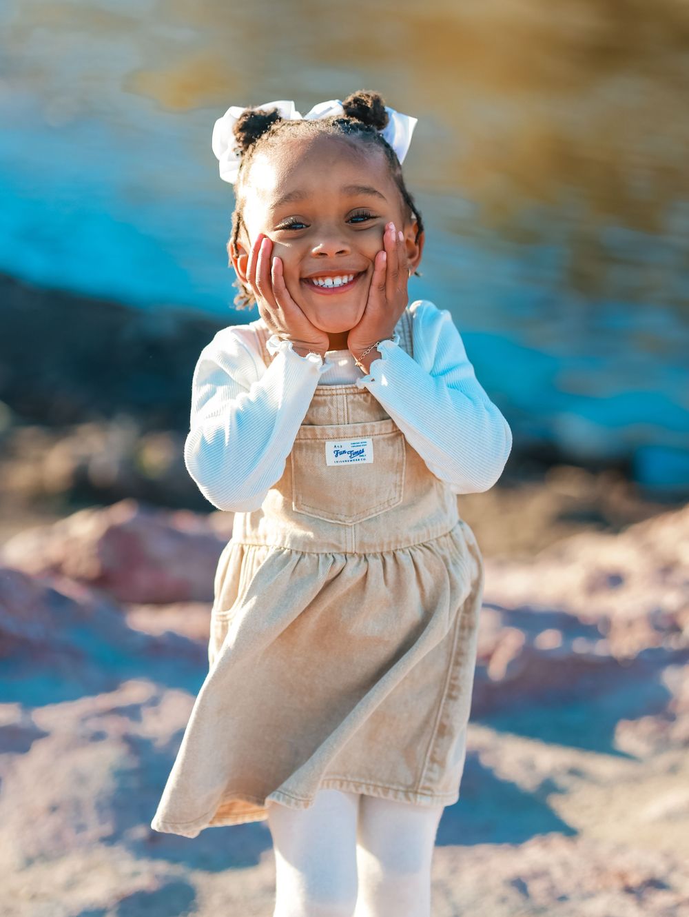 A child in a beige overall dress and white long sleeve shirt plays outdoors on a sunny autumn day.