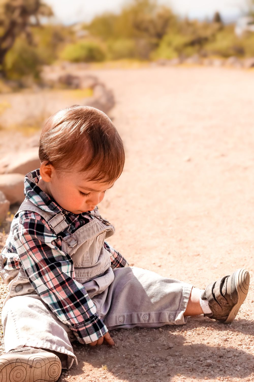 Small child in plaid shirt sitting on sandy ground outdoors.