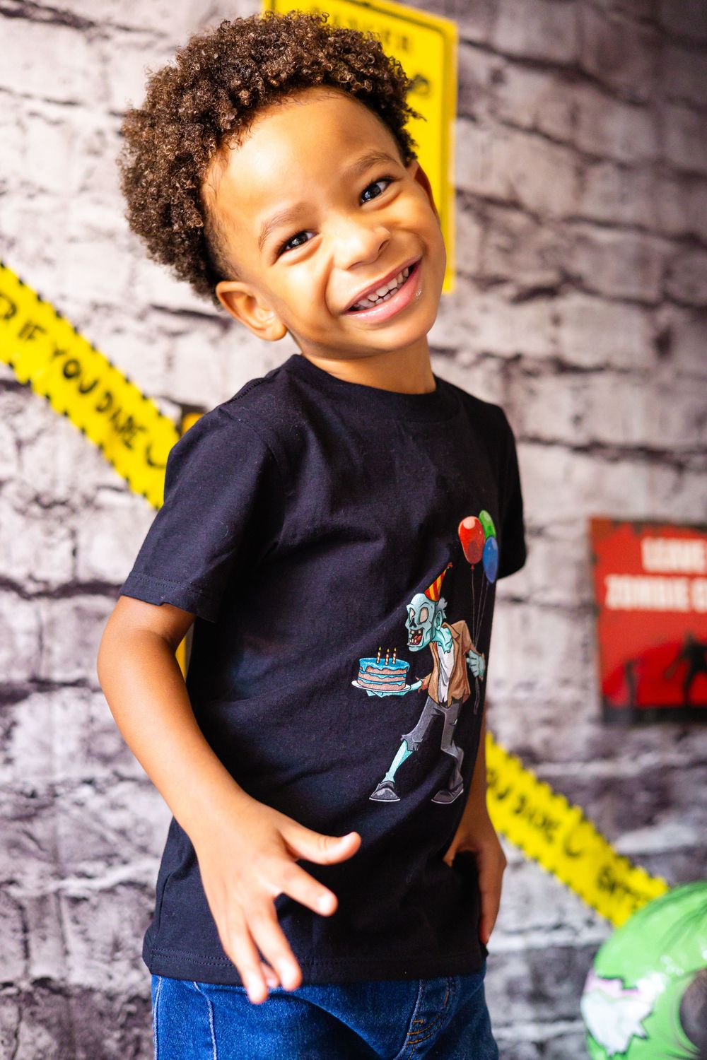 A young child wearing a navy t-shirt with a graphic design smiles and poses against a brick wall background.