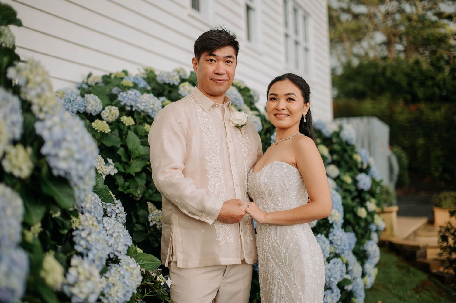 Couple poses in front of a wall decorated with blue hydrangeas at their wedding.