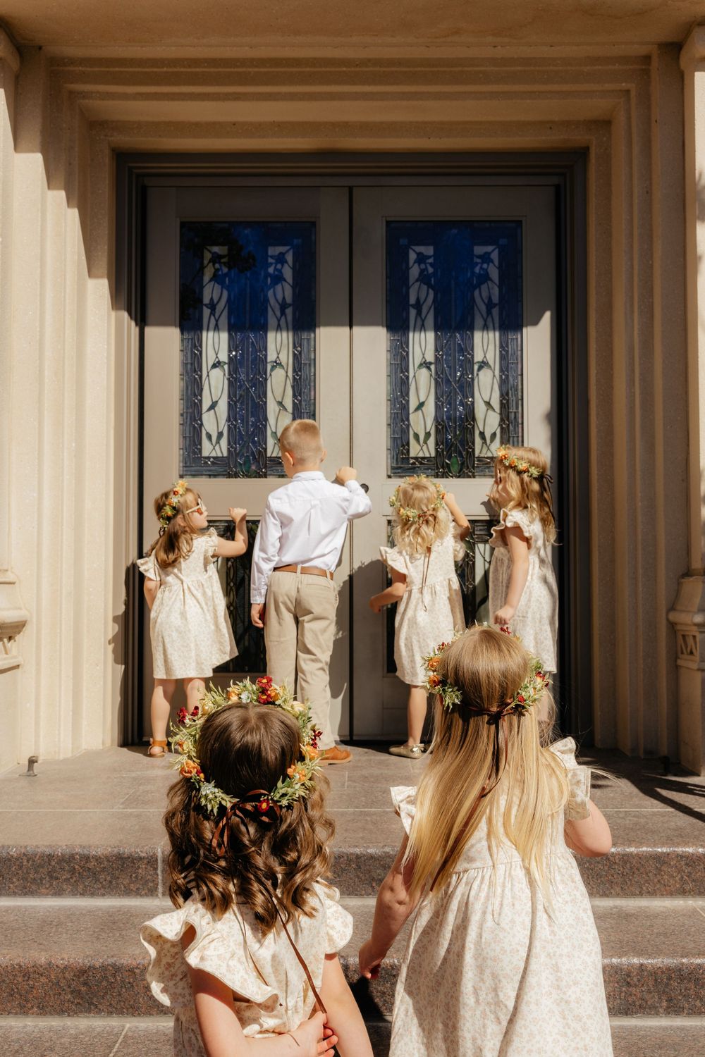 Children in flower crowns and white attire approach elegant temple doors on a sunny day.