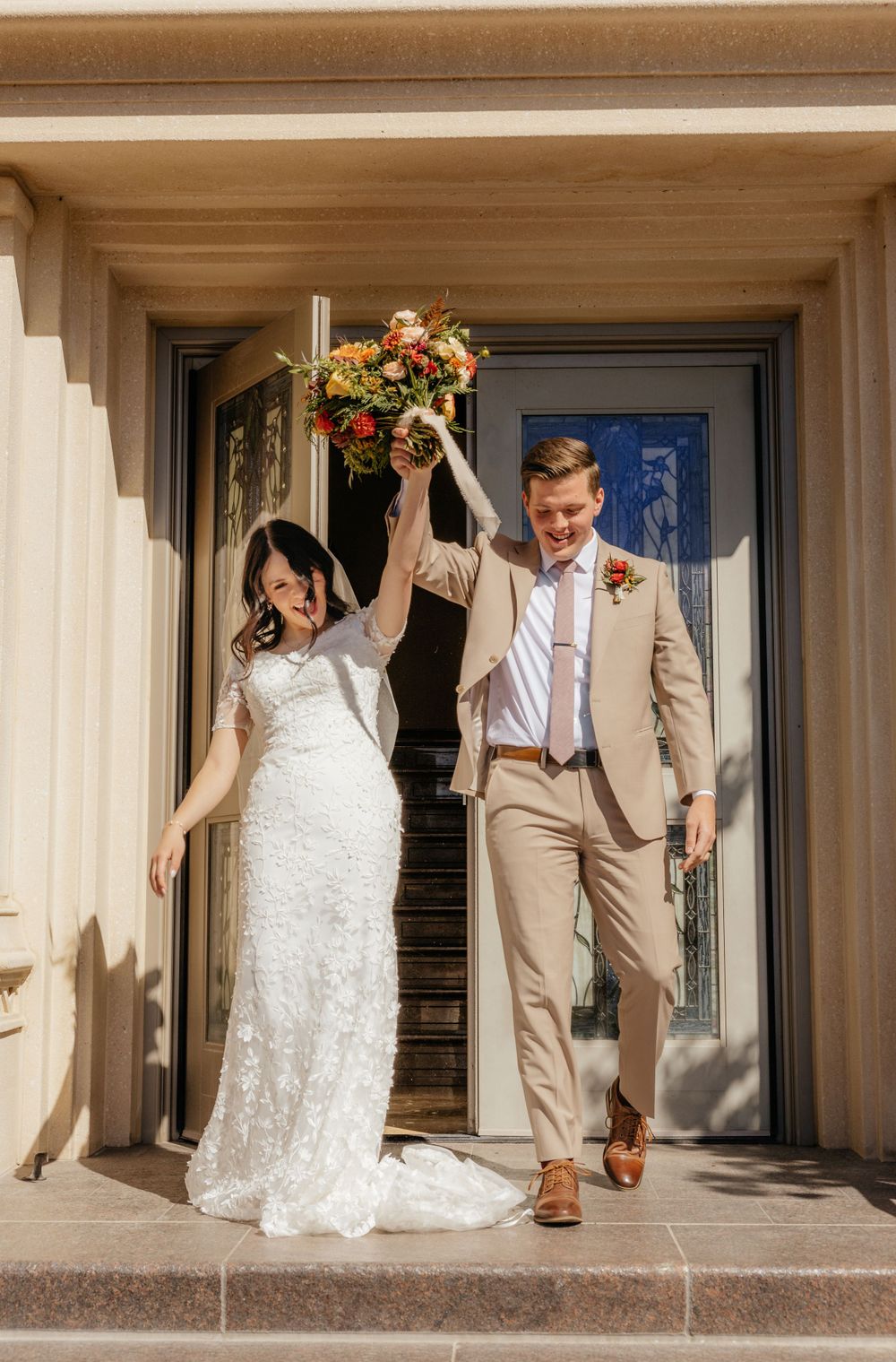 Smiling groom raises bride's bouquet triumphantly as couple exits temple after wedding.