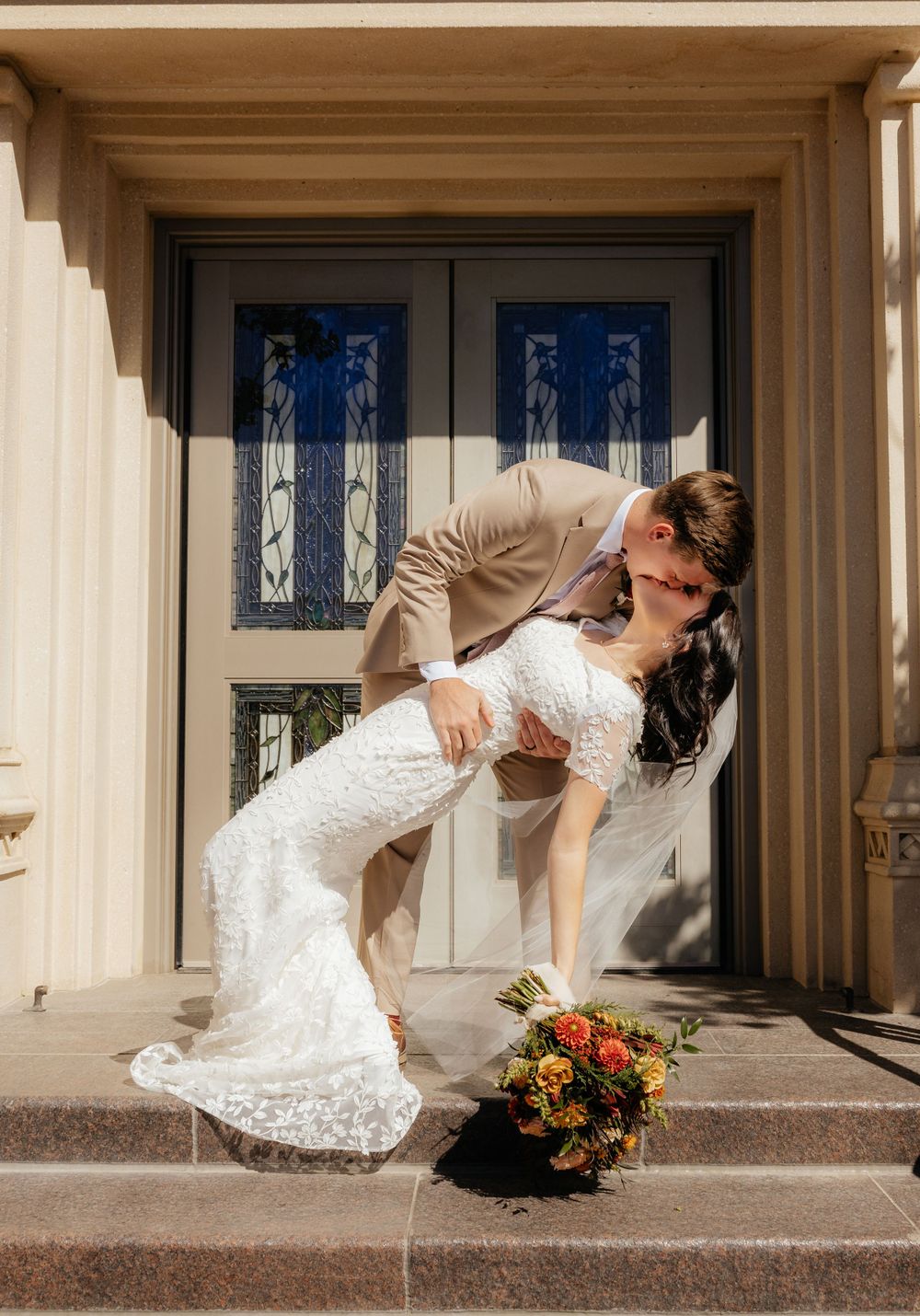 Groom dips bride for a romantic kiss on temple steps with colorful bouquet at their feet.