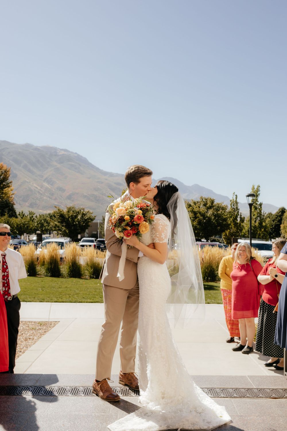 Bride and groom share first kiss outdoors with stunning mountain backdrop and guests watching.